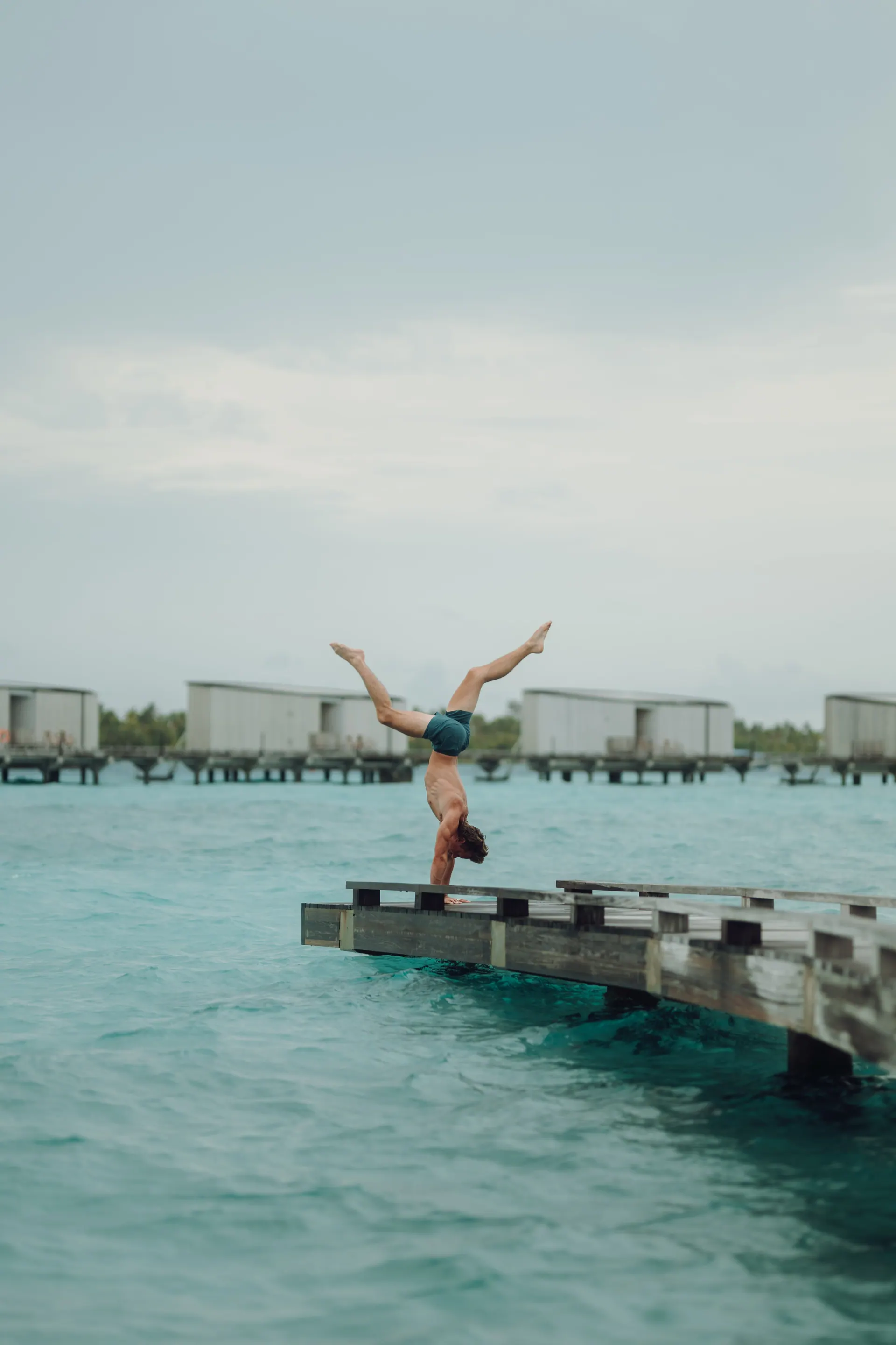 A yoga teacher holding a handstand on an overwater boardwalk above turquoise sea